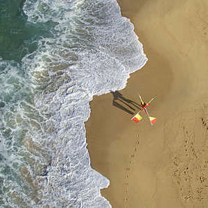 Areal view from a beach where you can see a lifeguard walking with one red and yellow safety flag on each shoulder crossing behind his neck. 