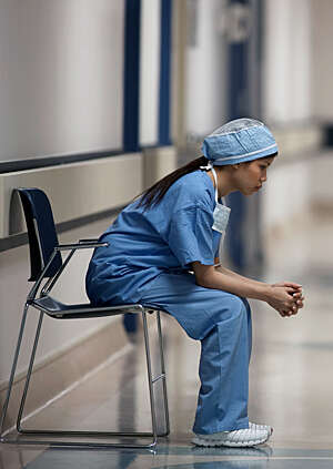 Female healthcare worker dressed in blue uniform sits on a chair lost in thought.