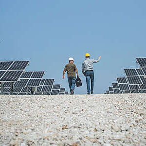 Back view of two technicians with safety helmets holding a work discussion as they walk across a field of solar panels set against bright blue skies.