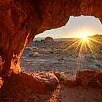 Dazzling ray of sun, seen through a red rock cavity, dances across a barren landscape at sunrise.