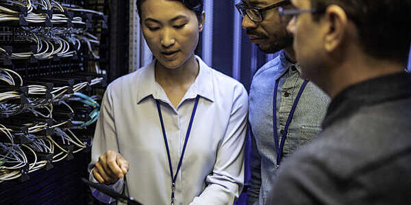 Three IT technician looking at a digital tablet and talking while standing next to a server in a data center.